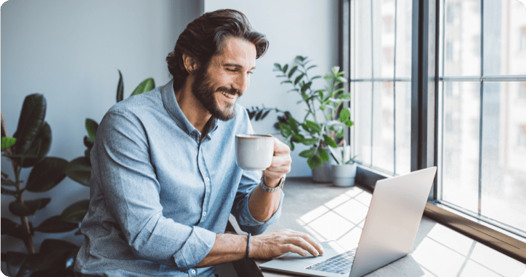 Photo d’un homme souriant qui consulte son ordinateur portable en prenant un café